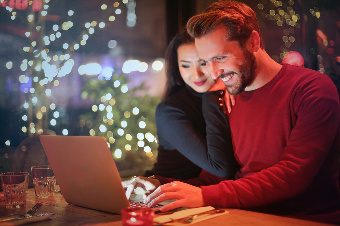 Photograph of a couple sitting closely together at a table, using a laptop in a dimly lit setting with colorful bokeh lights in the background. The scene conveys intimacy and collaboration, with the woman leaning on the man while he types on the keyboard.