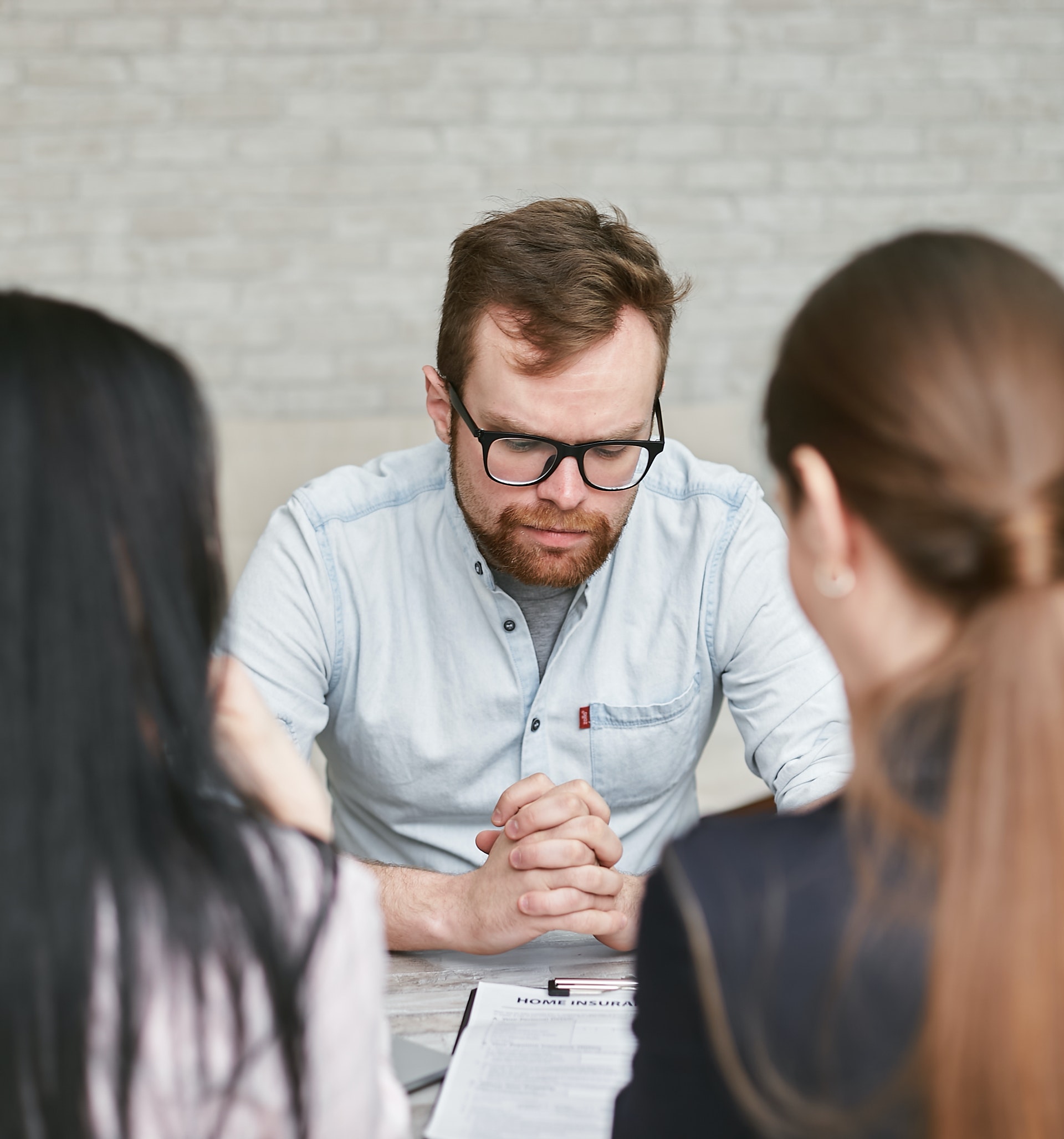Photograph of a man sitting at a table facing two women, likely engaged in a discussion or interview. Man wears a light blue shirt with hands clasped, while women are seen from behind, one with black hair and the other with brown hair tied back.