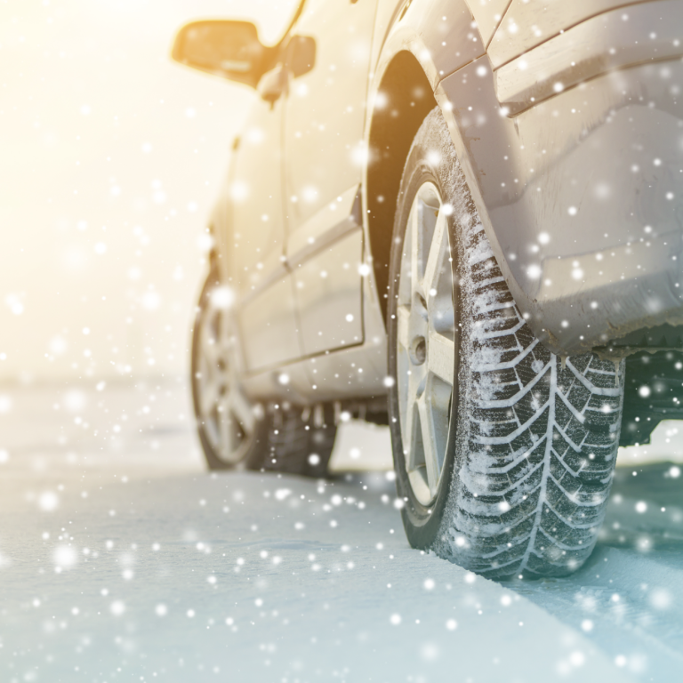 Photograph showing close-up of a car tire on snowy road with snowflakes falling and sunlight creating warm glow. Focus on tire tread designed for winter conditions highlights vehicle preparedness for icy, snowy driving.