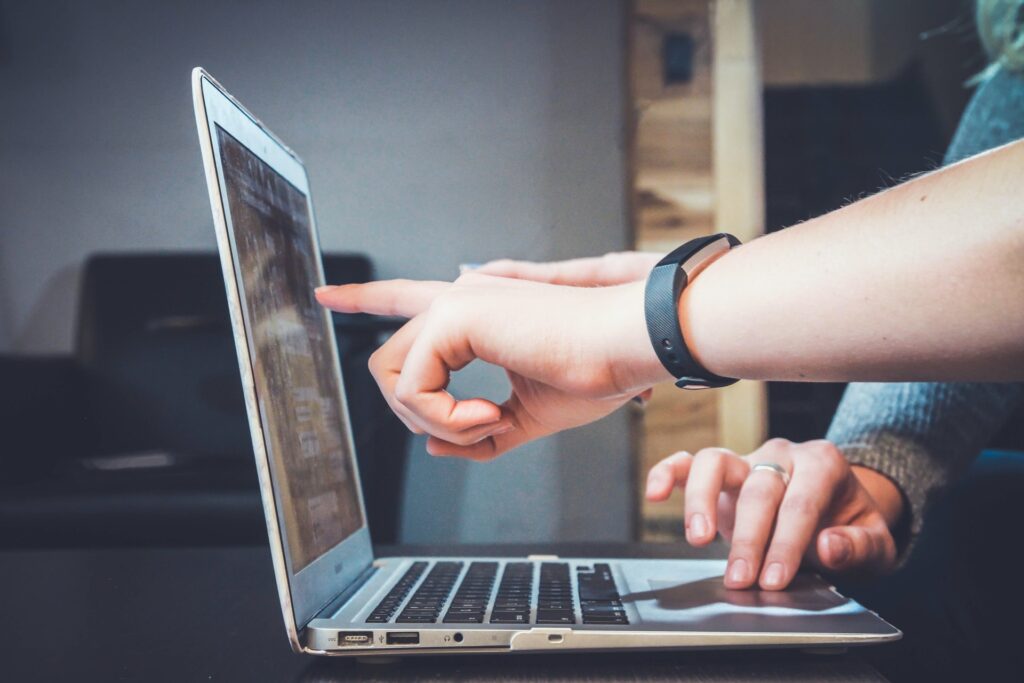 Photograph showing two people interacting with a laptop, one pointing at the screen while the other types on the keyboard. The setting appears to be an indoor workspace with a focus on collaboration or instruction.