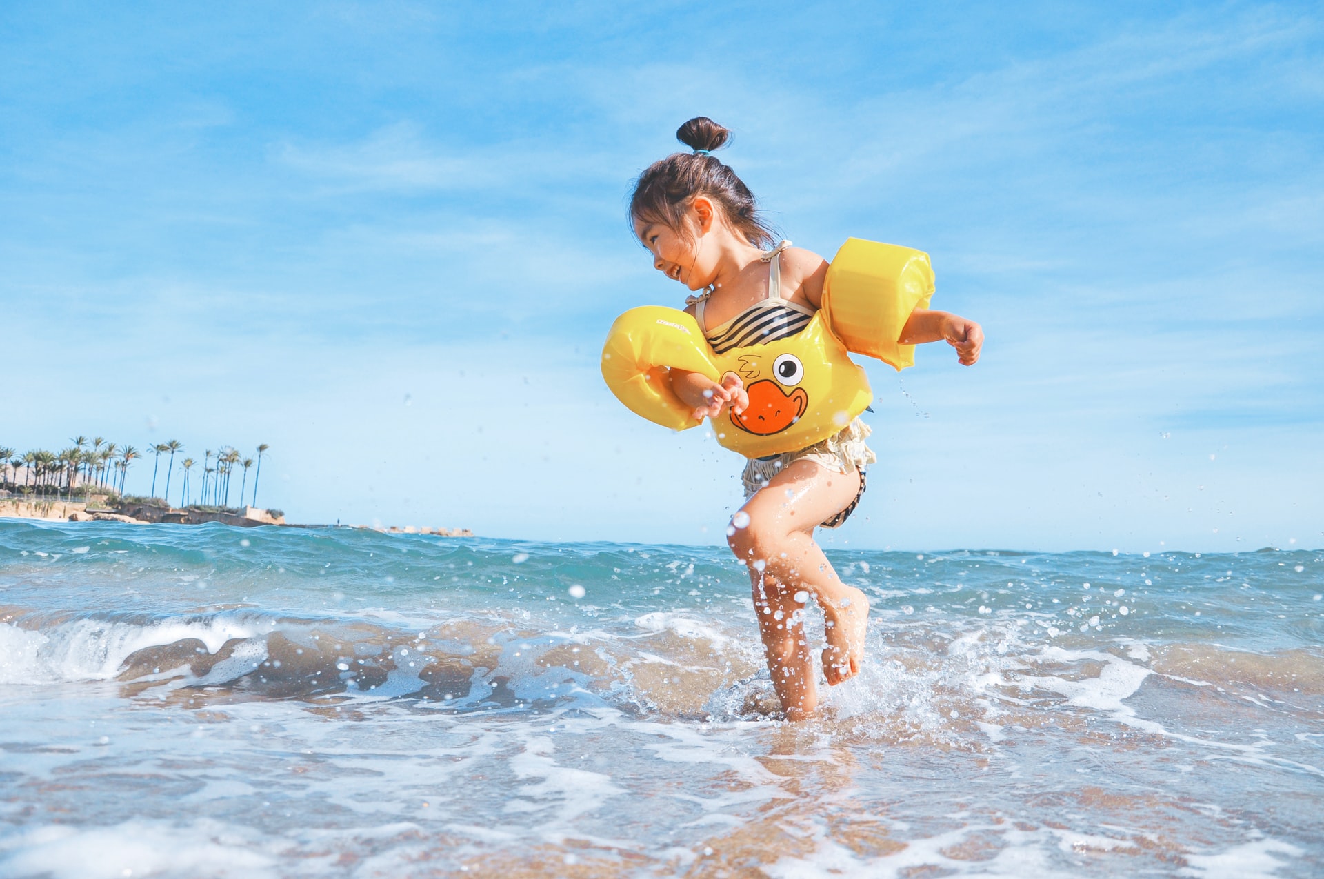 A joyful child plays in the surf, wearing yellow floaties and a duck-patterned swimsuit. Clear blue sky and palm trees in the background, conveying a sense of summer fun.