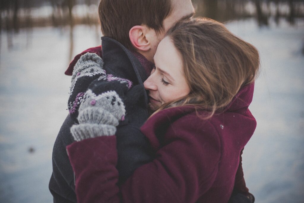 Photograph of two people embracing outdoors in a snowy setting, wearing winter clothing including a maroon coat and gray mittens with a cat design. The scene conveys warmth and affection against a cold, blurred background with trees.