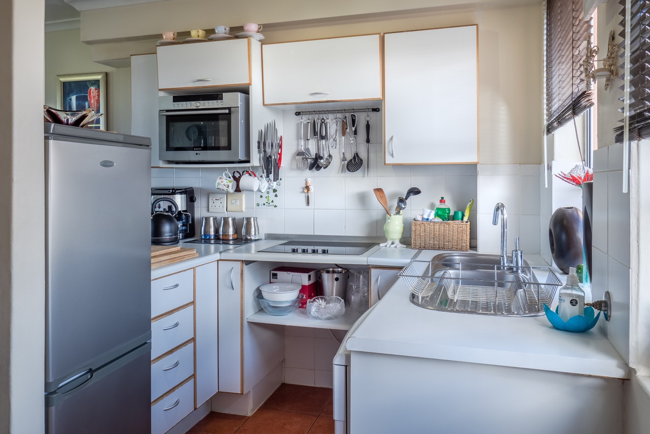 Photograph of a compact kitchen featuring white cabinets, a stainless steel refrigerator, and a built-in microwave. The countertop holds various kitchen utensils, a dish rack with drying dishes, and a basket with cleaning supplies, highlighting an organized and functional cooking space.