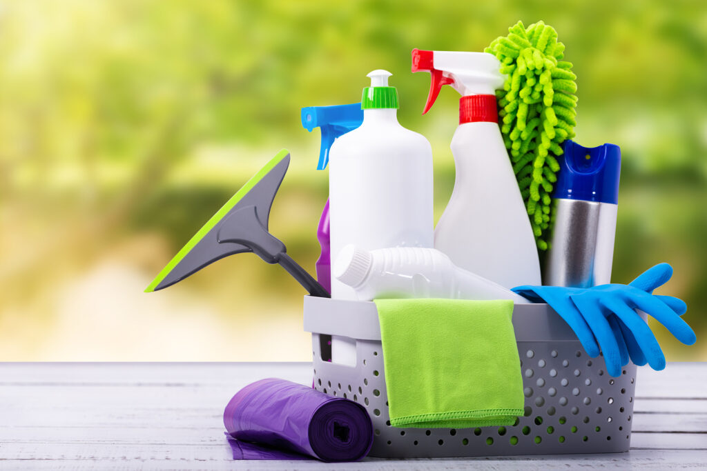 Photograph of cleaning supplies including sponges, a scrub brush, spray bottles, yellow rubber gloves, and a red basket against a blue background. Text in bold white letters asks, "Is Your Home Ready for Spring?" suggesting preparation for spring cleaning.