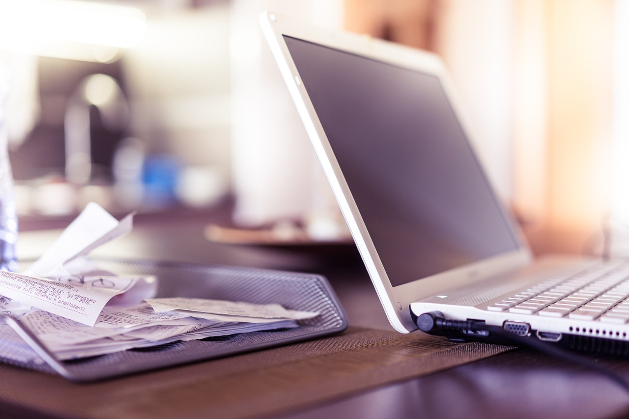 Photograph showing a close-up of a laptop on a table with a stack of receipts and papers beside it. The scene suggests a workspace or home office setting with soft natural lighting and a blurred background.