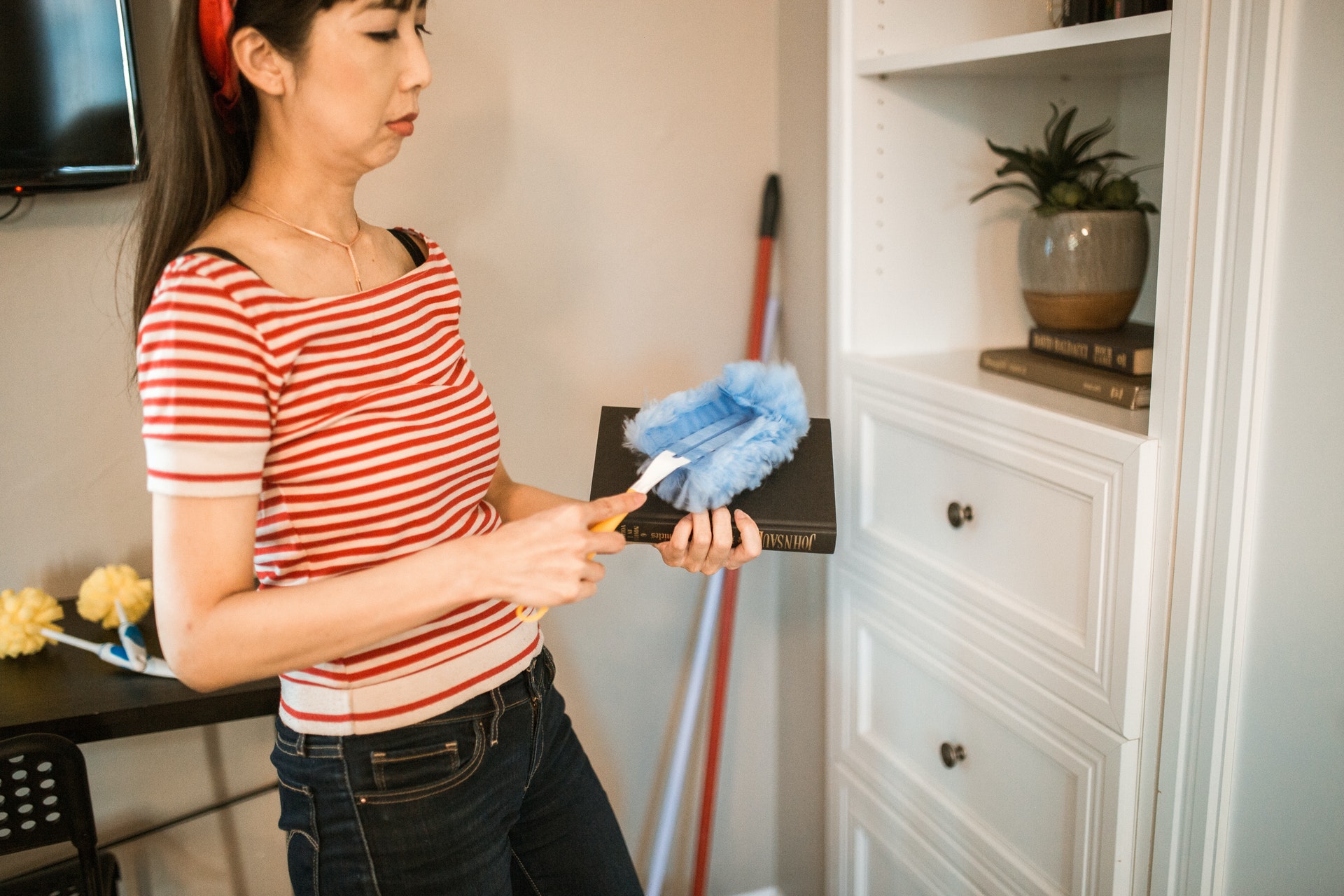 A woman in a red and white striped shirt and blue jeans is holding a dust cloth and cleaning a wooden shelf in a room with a plant and a lamp.