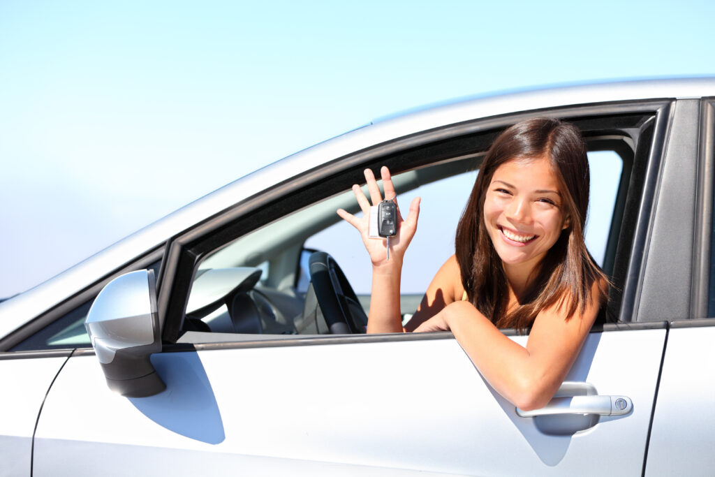 The image shows a smiling young person sitting behind the wheel of a car, holding up their left hand.