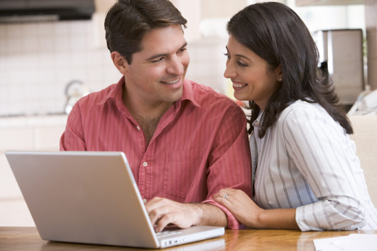 A couple is sitting at a kitchen table, intently working on a laptop surrounded by papers and documents.