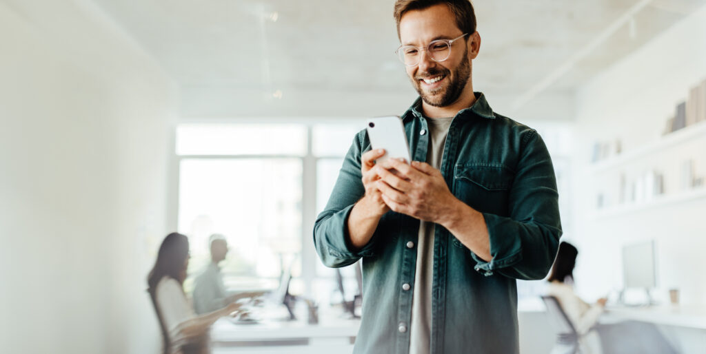 Photograph of a smiling man wearing glasses and a green shirt, looking at his smartphone in a bright office setting. Background shows blurred coworkers working at desks, highlighting a casual and productive work environment.