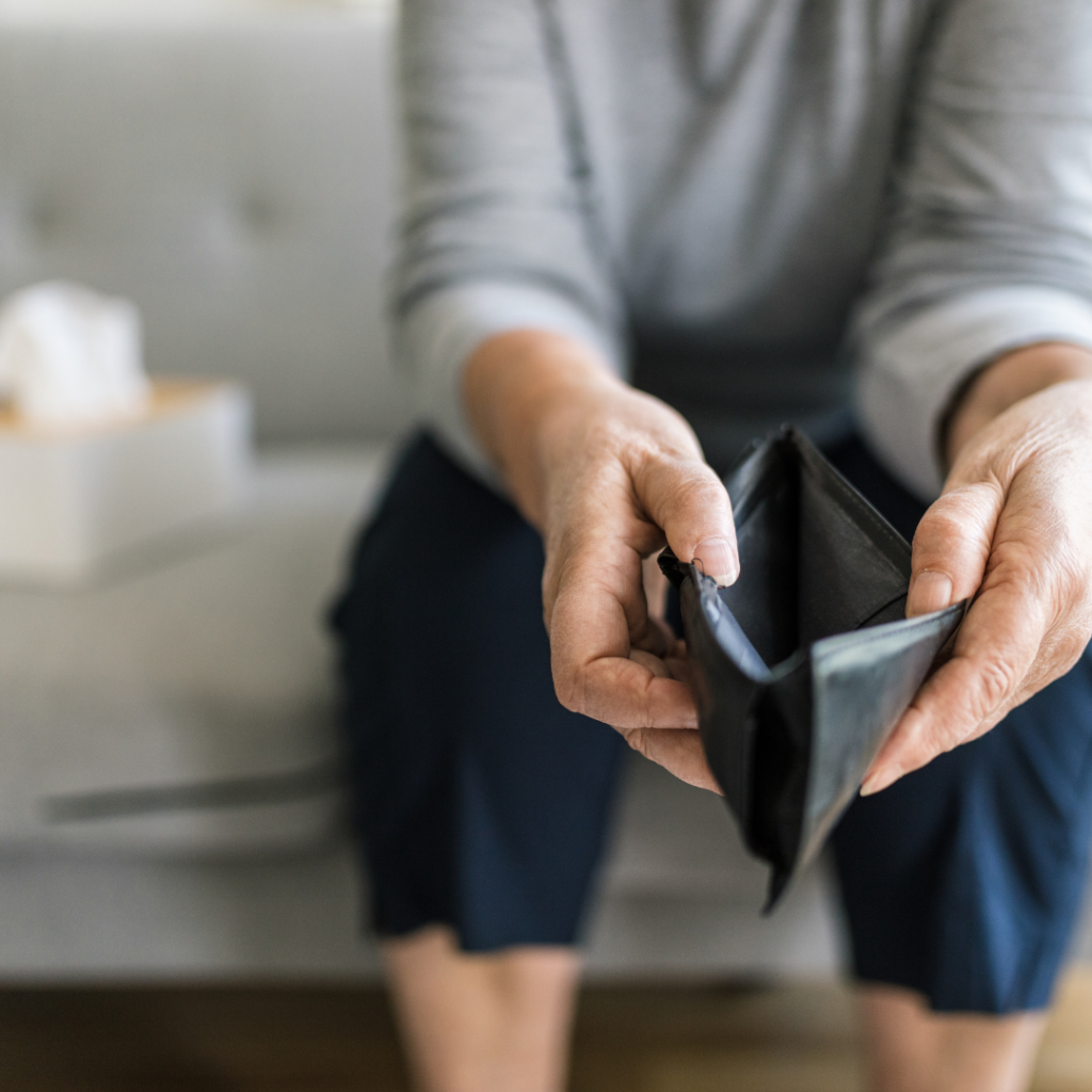 Photograph showing a person sitting on a couch holding an empty black wallet with both hands. The focus is on the wallet and hands, highlighting a theme of financial hardship or lack of money.