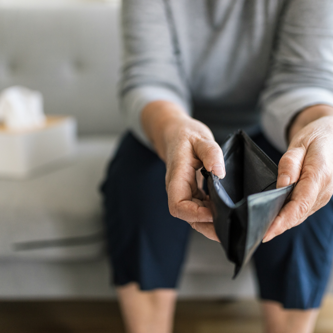 Photograph showing a person sitting on a couch holding an empty black wallet with both hands. The focus is on the wallet and hands, highlighting a theme of financial hardship or lack of money.